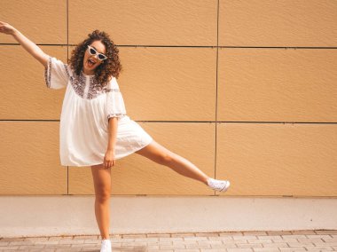 Yaz hipster beyaz elbise ve güneş gözlüğü giymiş afro bukleler saç modeli ile güzel gülümseyen model. Seksi kaygısız kız sarı duvara yakın sokakta poz. Komik ve pozitif kadın eğleniyor