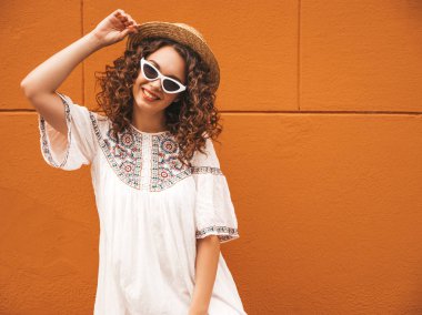 Yaz hipster beyaz elbise ve güneş gözlüğü giymiş afro bukleler saç modeli ile güzel gülümseyen model. Tasasız kız şapkaturuncu duvar ayakın sokakta poz. Komik ve pozitif kadın eğleniyor
