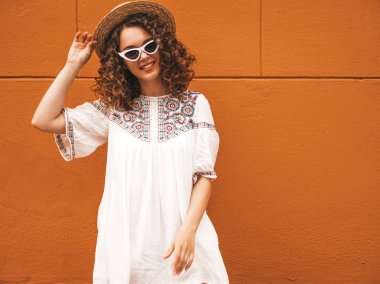 Yaz hipster beyaz elbise ve güneş gözlüğü giymiş afro bukleler saç modeli ile güzel gülümseyen model. Tasasız kız şapkaturuncu duvar ayakın sokakta poz. Komik ve pozitif kadın eğleniyor