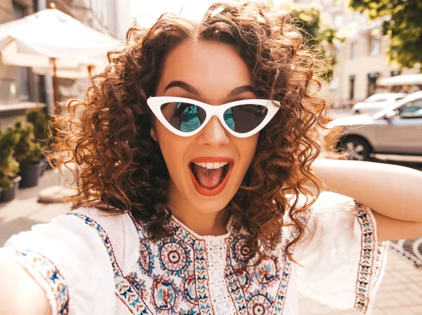 Beautiful smiling model with afro curls hairstyle dressed in summer hipster white dress.Sexy carefree girl posing in the street in sunglasses.Taking selfie self portrait photos on smartphone