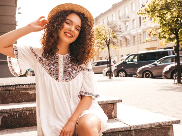 Beautiful smiling model with afro curls hairstyle dressed in summer hipster white dress.Sexy carefree girl posing in street.Trendy and positive woman having fun