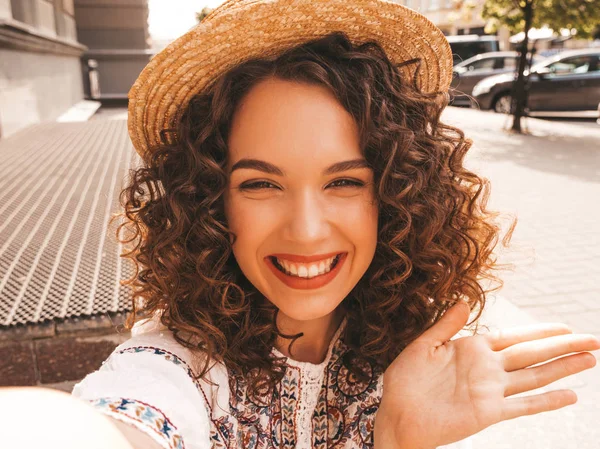 Beautiful smiling model with afro curls hairstyle dressed in summer hipster white dress.Sexy carefree girl posing in the street.Taking selfie self portrait photos on smartphone