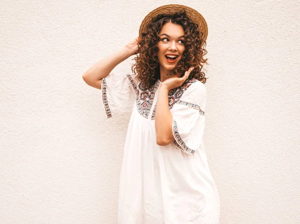 Beautiful smiling model with afro curls hairstyle dressed in summer hipster white dress.Sexy carefree girl posing in the street near white wall in hat.Funny, positive woman having fun and going crazy