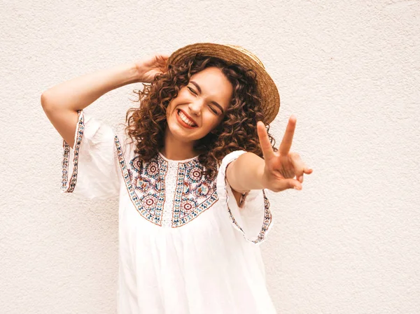 Beautiful smiling model with afro curls hairstyle dressed in summer hipster white dress.Sexy carefree girl posing in the street near white wall in hat.Funny, positive woman having fun and going crazy