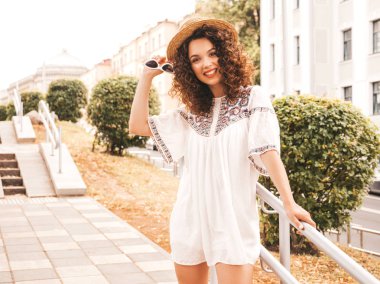 Yaz hipster beyaz elbise giymiş Afro bukleler saç ile güzel gülümseyen model. Seksi kaygısız kız sokakta poz. Şık komik ve pozitif kadın güneş gözlüğü ve şapka eğlenmek