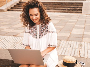Yaz hipster beyaz elbise giymiş afro bukleler saç modeli ile güzel gülümseyen model. Sokakta oturan seksi kaygısız kız. Dizüstü bilgisayar kullanan modaya uygun kadın. Dizüstü bilgisayarda bilgi yazma ve arama