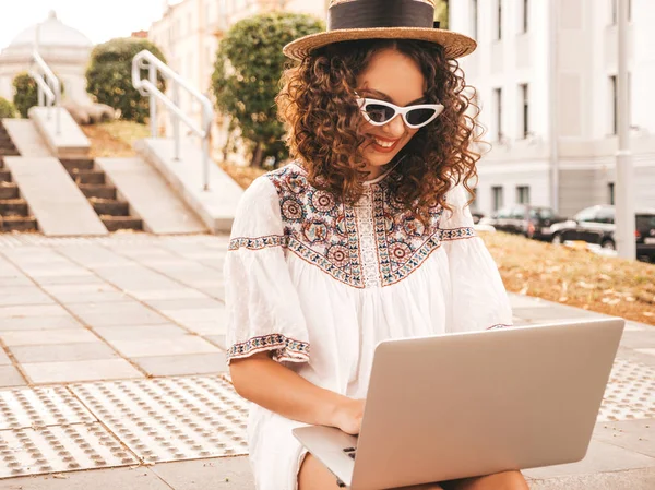 Beautiful smiling model with afro curls hairstyle in summer hipster white dress and hat.Sexy carefree girl sitting in the street.Trendy woman using notebook.Typing and searching information in laptop