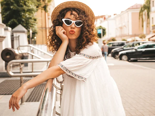 Beautiful smiling model with afro curls hairstyle dressed in summer hipster white dress.Sexy carefree girl posing in street.Positive woman having fun in sunglasses and hat.Makes duck face