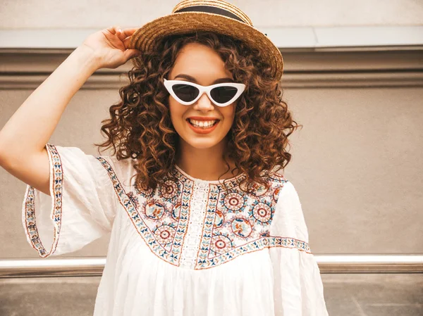 Beautiful smiling model with afro curls hairstyle dressed in summer hipster white dress.Sexy carefree girl posing in street.Trendy funny and positive woman having fun in sunglasses and hat