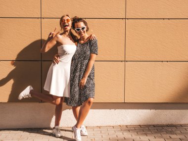 Trendy yaz elbisesi iki genç güzel gülümseyen hipster kız. Seksi kaygısız kadınlar sarı duvarın yakınındaki sokakta poz. Güneş gözlüğü ile eğlenen pozitif modeller. Sarılma, barış işareti