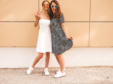 Moda yazlık elbiseler iki genç güzel gülümseyen hipster kız. Seksi kaygısız kadınlar sarı duvarın yakınındaki sokakta poz. Güneş gözlüğü ile eğlenen pozitif modeller