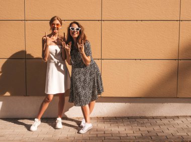 Trendy yaz elbisesi iki genç güzel gülümseyen hipster kız. Seksi kaygısız kadınlar sarı duvarın yakınındaki sokakta poz. Güneş gözlüğü ile eğlenen pozitif modeller. Sarılma, dil ve barış işareti gösterir