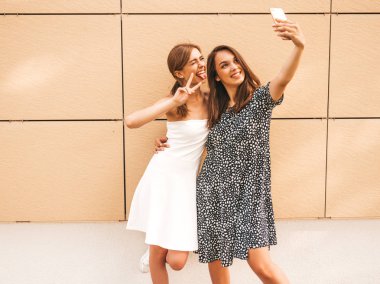 Yazlık kıyafetler giyen iki genç gülümseyen hipster kadın. Akıllı telefonda selfie selfie portre fotoğrafları çeken kızlar. Sarı duvarın yanındaki sokak arka planında poz veren mankenler. Kadın olumlu yüz duyguları gösteren