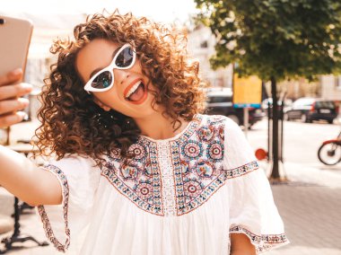 Yaz hipster beyaz elbise giymiş afro bukleler saç modeli ile güzel gülümseyen model. Seksi kaygısız kız güneş gözlüğü sokakta poz. Akıllı telefonda selfie selfie portre fotoğrafları çekmek