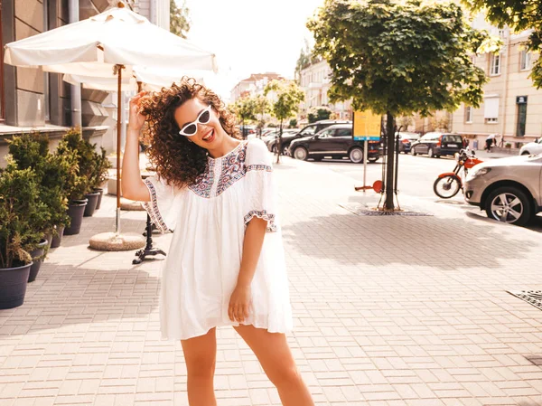 Beautiful smiling model with afro curls hairstyle dressed in summer hipster white dress.Sexy carefree girl posing in the street background.Trendy funny and positive woman having fun in sunglasses