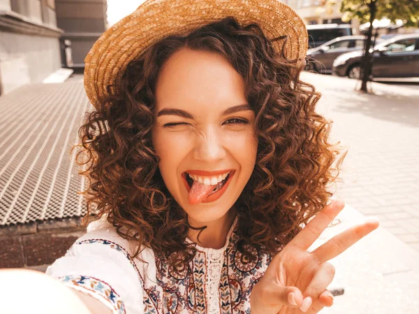 Beautiful smiling model with afro curls hairstyle dressed in summer hipster white dress.Sexy carefree girl posing in the street.Taking selfie self portrait photos in hat on smartphone.Shows tongue