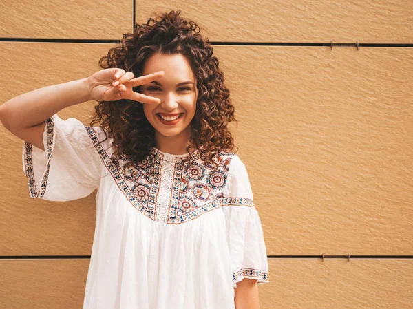 Beautiful smiling model with afro curls hairstyle dressed in summer hipster white dress.Sexy carefree girl posing in the street near yellow wall.Funny and positive woman having fun.Shows peace sign