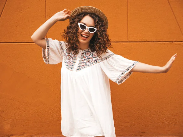 Beautiful smiling model with afro curls hairstyle dressed in summer hipster white dress and sunglasses.Carefree girl posing in the street near orange wall in hat.Funny and positive woman having fun
