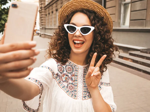 Beautiful smiling model with afro curls hairstyle dressed in summer hipster white dress.Sexy carefree girl posing in the street in sunglasses.Taking selfie self portrait photos in hat on smartphone