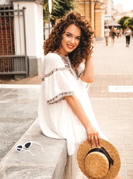 Beautiful smiling model with afro curls hairstyle dressed in summer hipster white dress.Sexy carefree girl posing in street.Trendy funny and positive woman having fun in hat