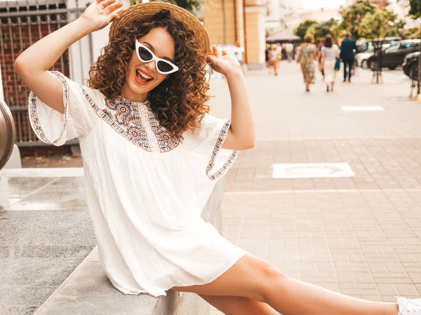 Beautiful smiling model with afro curls hairstyle dressed in summer hipster white dress.Sexy carefree girl posing in street.Trendy funny and positive woman having fun in sunglasses and hat