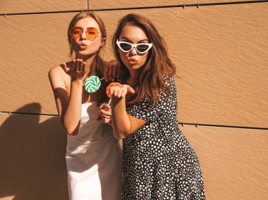 Trendy yaz elbisesi iki genç güzel gülümseyen hipster kız. Seksi kaygısız kadınlar sarı duvarın yakınındaki sokakta poz. Güneş gözlüğü ile eğlenen pozitif modeller. Şeker lolipop yeme