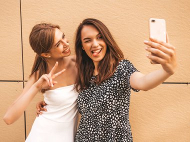Yazlık kıyafetler giyen iki genç gülümseyen hipster kadın. Akıllı telefonda selfie selfie portre fotoğrafları çeken kızlar. Sarı duvarın yanındaki sokak arka planında poz veren mankenler. Kadın olumlu yüz duyguları gösteren