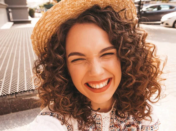 Beautiful smiling model with afro curls hairstyle dressed in summer hipster white dress.Sexy carefree girl posing in the street.Taking selfie self portrait photos in hat on smartphone