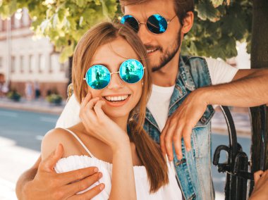 Gülümseyen güzel kız ve yakışıklı erkek arkadaşı. Gündelik yazlık elbiseli kadın ve kot elbiseli adam. Mutlu, neşeli bir aile. Kadın arka planda eğleniyor. Güneş gözlüklü çifte sarılıyor.