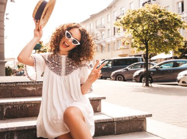 Yaz hipster beyaz elbise giymiş afro bukleler saç modeli ile güzel gülümseyen model. Seksi kaygısız kız sokakta poz. Trendy ve pozitif kadın güneş gözlüğü ve şapka eğlenmek. Barış işaretini gösterir