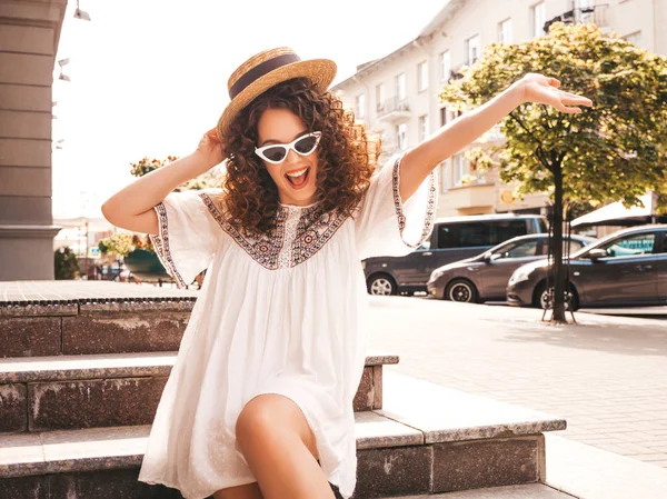 Beautiful smiling model with afro curls hairstyle dressed in summer hipster white dress.Sexy carefree girl posing in street.Trendy and positive woman having fun in sunglasses and hat