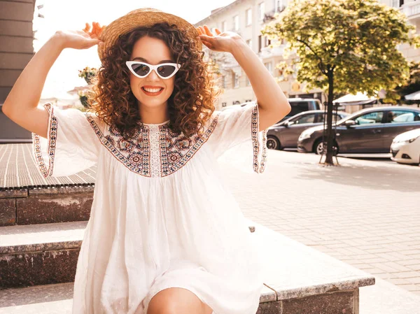 Beautiful smiling model with afro curls hairstyle dressed in summer hipster white dress.Sexy carefree girl posing in street.Trendy and positive woman having fun in sunglasses and hat