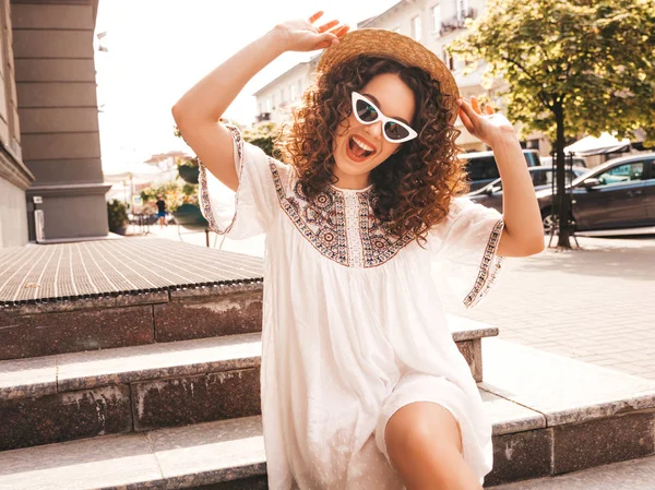 Beautiful smiling model with afro curls hairstyle dressed in summer hipster white dress.Sexy carefree girl posing in street.Trendy and positive woman having fun in sunglasses and hat