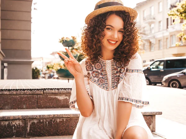 Beautiful smiling model with afro curls hairstyle dressed in summer hipster white dress.Sexy carefree girl posing in street.Trendy and positive woman having fun in hat.Shows peace sign