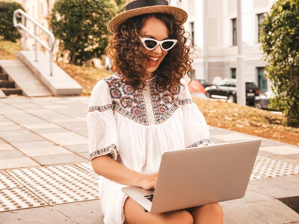 Beautiful smiling model with afro curls hairstyle dressed in summer hipster white dress.Sexy carefree girl sitting in the street.Trendy woman using notebook.Typing and searching information in laptop