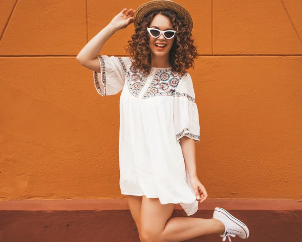 Beautiful smiling model with afro curls hairstyle dressed in summer hipster white dress and sunglasses.Carefree girl posing in the street near orange wall in hat.Funny and positive woman having fun