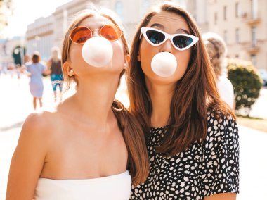 Moda yazlık giysiler iki genç güzel gülümseyen hipster kız Portresi. Sokak arka planında poz veren kaygısız kadınlar. Güneş gözlüğü ve sakız ile kabarcık üfleme eğlenmek olumlu modeller
