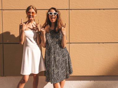 Trendy yaz elbisesi iki genç güzel gülümseyen hipster kız. Seksi kaygısız kadınlar sarı duvarın yakınındaki sokakta poz. Güneş gözlüğü ile eğlenen pozitif modeller. Sarılma, rock and roll işareti gösterir