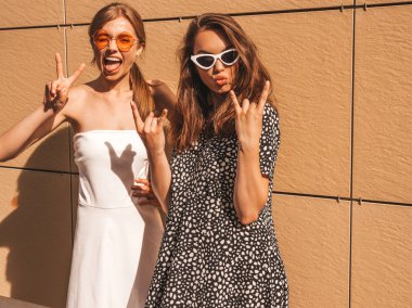 Trendy yaz elbisesi iki genç güzel gülümseyen hipster kız. Seksi kaygısız kadınlar sarı duvarın yakınındaki sokakta poz. Güneş gözlüğü ile eğlenen pozitif modeller. Sarılma, rock and roll işareti gösterir