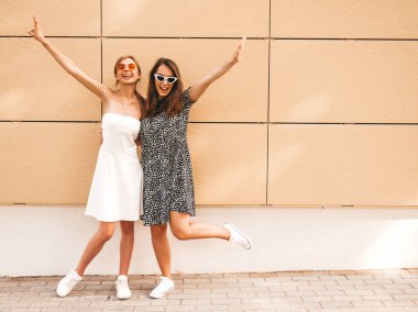 Moda yazlık elbiseler iki genç güzel gülümseyen hipster kız. Seksi kaygısız kadınlar sarı duvarın yakınındaki sokakta poz. Güneş gözlüğü ile eğlenen pozitif modeller. El kaldırma