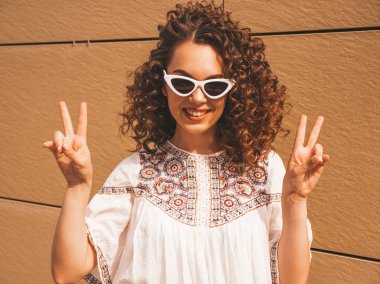 Yaz hipster beyaz elbise ve güneş gözlüğü giymiş afro bukleler saç modeli ile güzel gülümseyen model. Seksi kaygısız kız sarı duvara yakın sokakta poz. Komik kadın barış işareti gösterir
