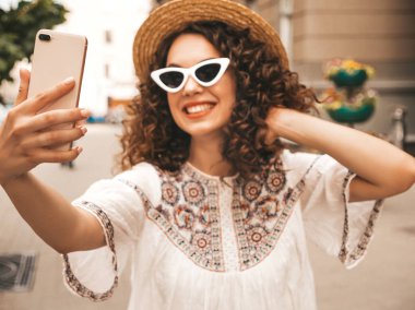 Yaz hipster beyaz elbise giymiş afro bukleler saç modeli ile güzel gülümseyen model. Seksi kaygısız kız güneş gözlüğü sokakta poz. Şapkalı akıllı telefonda selfie selfie portre fotoğrafları çekmek