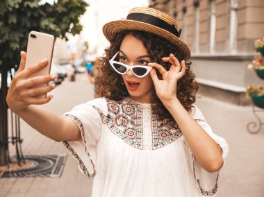 Yaz hipster beyaz elbise giymiş afro bukleler saç modeli ile güzel gülümseyen model. Seksi kaygısız kız güneş gözlüğü sokakta poz. Akıllı telefonda selfie selfie portre fotoğrafları çekmek