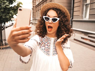 Yaz hipster elbise giymiş afro bukleler saç modeli ile güzel gülümseyen model. Sokakta güneş gözlüğüyle poz veren bir kız. Akıllı telefonda şapkayla selfie selfie portre fotoğrafları çekmek. Şok ve şaşırttı