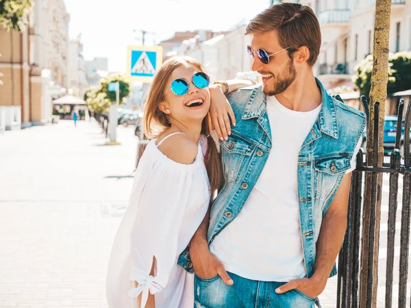 Gülümseyen güzel kız ve onun yakışıklı erkek arkadaşı sokakta poz. Rahat yazlık elbiseli kadın ve kot pantolon giyen bir erkek. Mutlu neşeli çift aile güneş gözlüğü eğlenmek