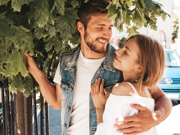 Gülümseyen güzel kız ve onun yakışıklı erkek arkadaşı ağacın yanında sokakta poz. Rahat yazlık elbiseli kadın ve kot pantolon giyen bir erkek. Mutlu neşeli çift aile eğleniyor