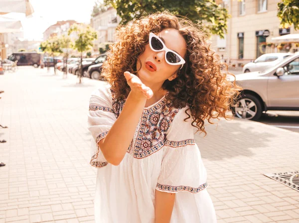 Beautiful smiling model with afro curls hairstyle dressed in summer hipster white dress.Sexy carefree girl posing in street.Trendy funny and positive woman having fun in sunglasses.Gives air kiss