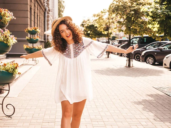 Beautiful smiling model with afro curls hairstyle dressed in summer hipster white dress. Sexy carefree girl posing in the street background. Trendy funny and positive woman having fun.Raising hands