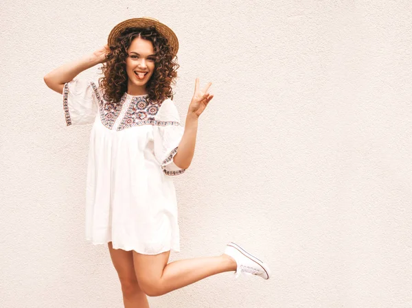 Beautiful smiling model with afro curls hairstyle dressed in summer hipster white dress.Sexy carefree girl posing in the street near white wall in hat.Funny, positive woman having fun and going crazy