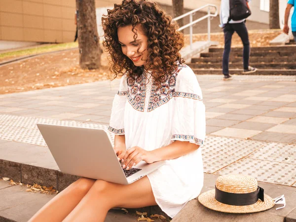 Beautiful smiling model with afro curls hairstyle dressed in summer hipster white dress.Sexy carefree girl sitting in the street.Trendy woman using notebook.Typing and searching information in laptop